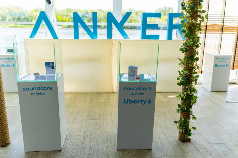 Produkty marki Anker wyeksponowane w szklanych gablotach wystawowych, na białych postumentach z nazwami modeli, w tle duże niebieskie litery tworzące napis Anker, po bokach dekoracyjne rośliny i okna z widokiem na rzekę.