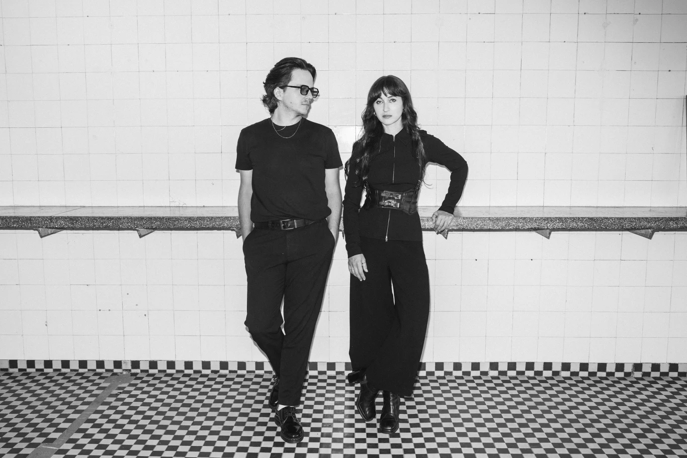 Blauka to Piotr Lewańczyk i Georgina Tarasiuk Dwie osoby ubrane na czarno stoją w nowoczesnym, kafelkowanym pomieszczeniu z czarno-białą podłogą, opierając się o blat i patrząc w stronę aparatu.