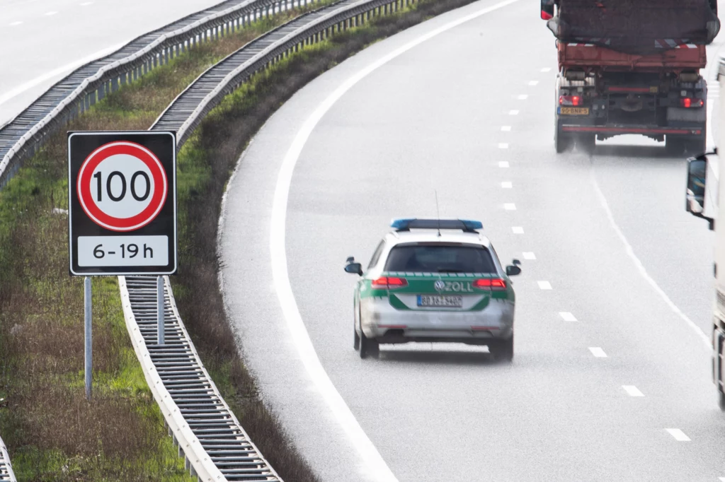 Zniesienie ograniczenia prędkości do 100 km/h zostanie zniesione początkowo na trzech odcinkach autostrad. Samochód policyjny oraz ciężarówki jadące autostradą, po lewej stronie widoczny znak drogowy ograniczający prędkość do 100 km/h w godzinach od 6 do 19, asfaltowa jezdnia na łuku drogi, barierki ochronne wzdłuż jezdni.