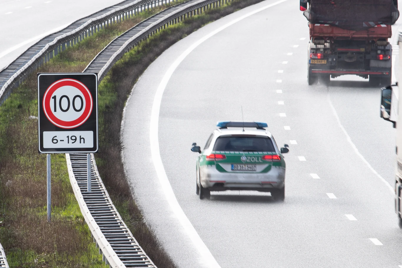 Zniesienie ograniczenia prędkości do 100 km/h zostanie zniesione początkowo na trzech odcinkach autostrad. Samochód policyjny oraz ciężarówki jadące autostradą, po lewej stronie widoczny znak drogowy ograniczający prędkość do 100 km/h w godzinach od 6 do 19, asfaltowa jezdnia na łuku drogi, barierki ochronne wzdłuż jezdni.