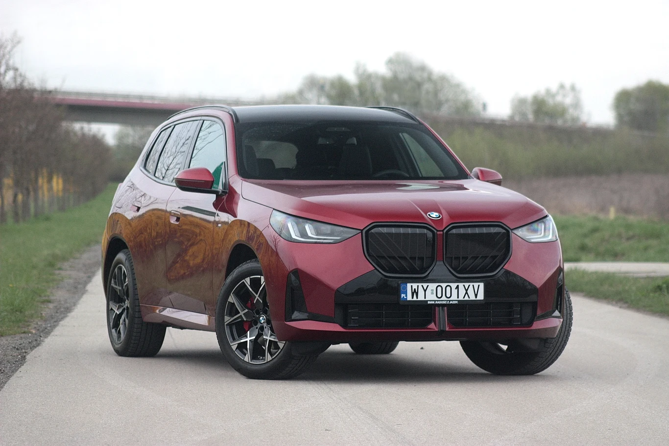 Najpopularniejszym samochodem BMW w Polsce w 2025 roku było X3. Czerwony samochód typu SUV marki BMW stoi na asfaltowej drodze, z przodu widoczne czarne osłony chłodnicy oraz tablica rejestracyjna z numerem WY-001XV, w tle drzewa i most.