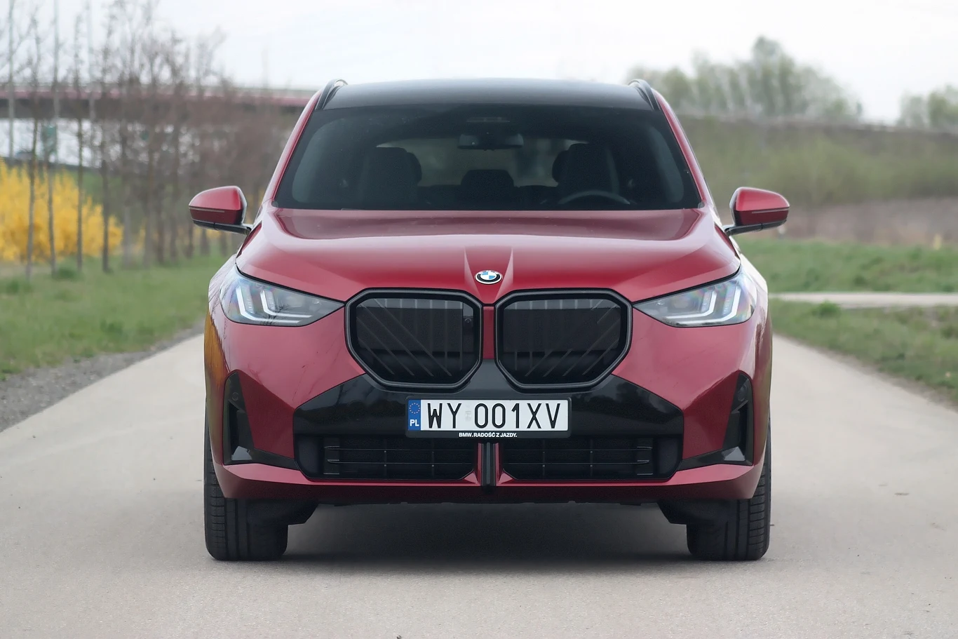 Czerwony samochód marki BMW na środku asfaltowej drogi, widoczny z przodu z czytelnymi światłami i tablicą rejestracyjną. W tle rozmyte elementy przyrody oraz szosa.