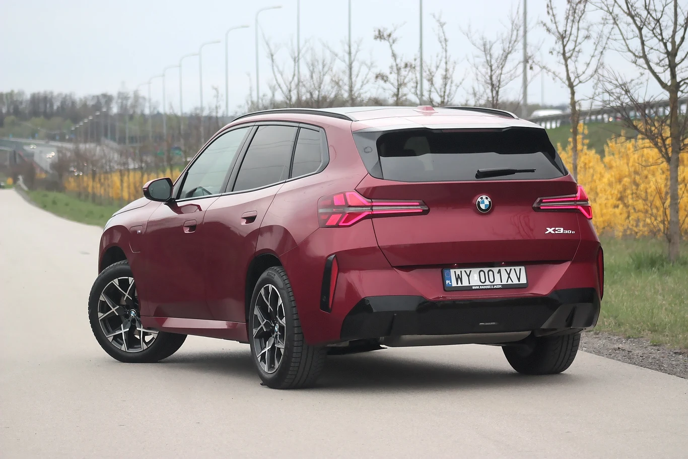 Czerwony SUV BMW iX stojący na asfaltowej drodze, widok od tyłu z wyraźnie widoczną tablicą rejestracyjną, w tle roślinność i szpaler drzew.