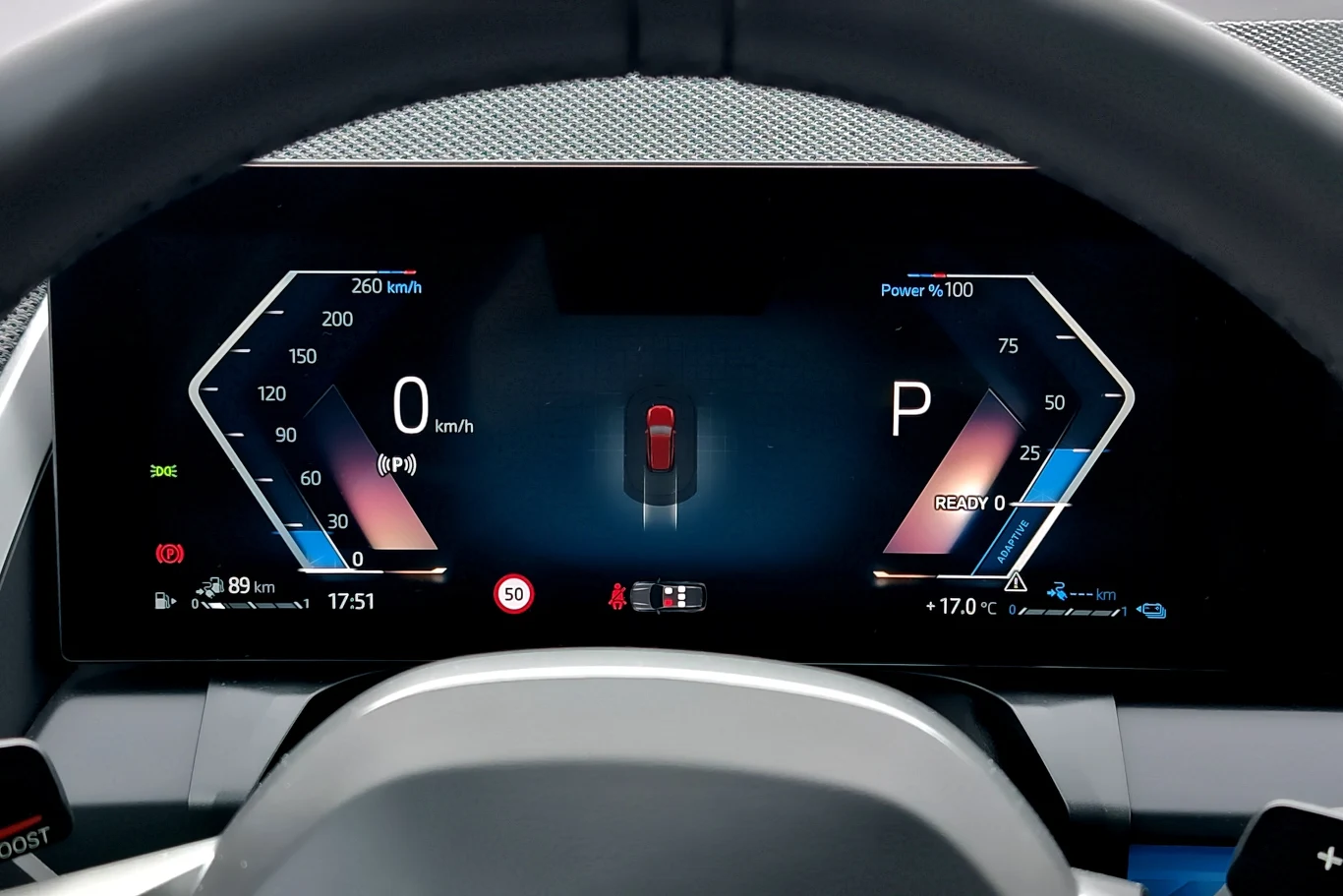 Cyfrowy kokpit samochodu z elektronicznym wskaźnikiem prędkości i obrotomierzem, trybem jazdy P oraz graficznym przedstawieniem auta pośrodku ekranu. Kierownica widoczna w dolnej części kadru.