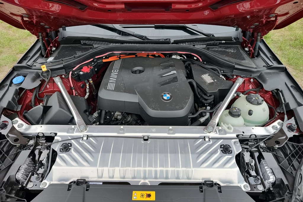 W nowym hybrydowym układzie napędowym BMW X3 rozbudowano część elektryczną Silnik samochodu marki BMW umieszczony pod otwartą maską, widoczne elementy konstrukcyjne komory silnika, instalacje i obudowa jednostki napędowej, dominują odcienie czerni, metalu oraz czerwonego lakieru karoserii.