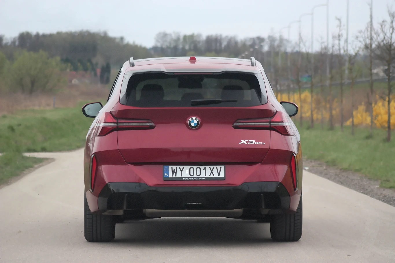Czerwony samochód BMW iX3 stojący na pustej asfaltowej drodze, widoczny od tyłu, wyraźnie widoczna tablica rejestracyjna i emblematy modelu, w tle drzewa i pola.