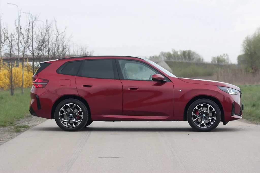 BMW X3 Bordowy samochód typu SUV zaparkowany na betonowej drodze w otoczeniu wiosennej zieleni i krzewów, widoczny z profilu, posiada nowoczesne reflektory i czarne szyby.