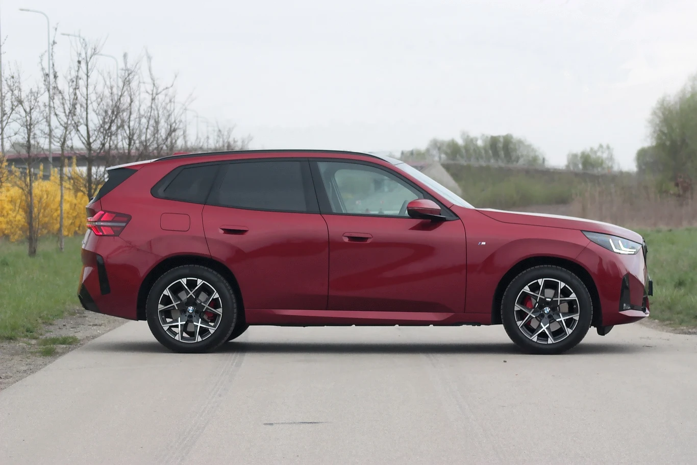 Bordowy samochód typu SUV zaparkowany na betonowej drodze w otoczeniu wiosennej zieleni i krzewów, widoczny z profilu, posiada nowoczesne reflektory i czarne szyby.