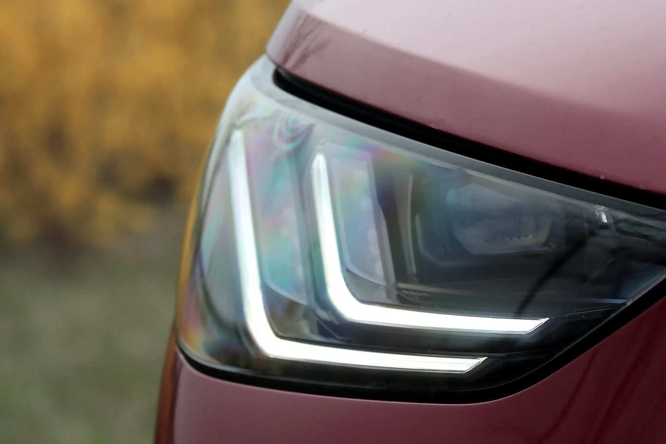 Zbliżenie na nowoczesny reflektor LED samochodu o bordowym kolorze nadwozia, z wyraźnie zaznaczonymi liniami świetlnymi w kształcie litery L.