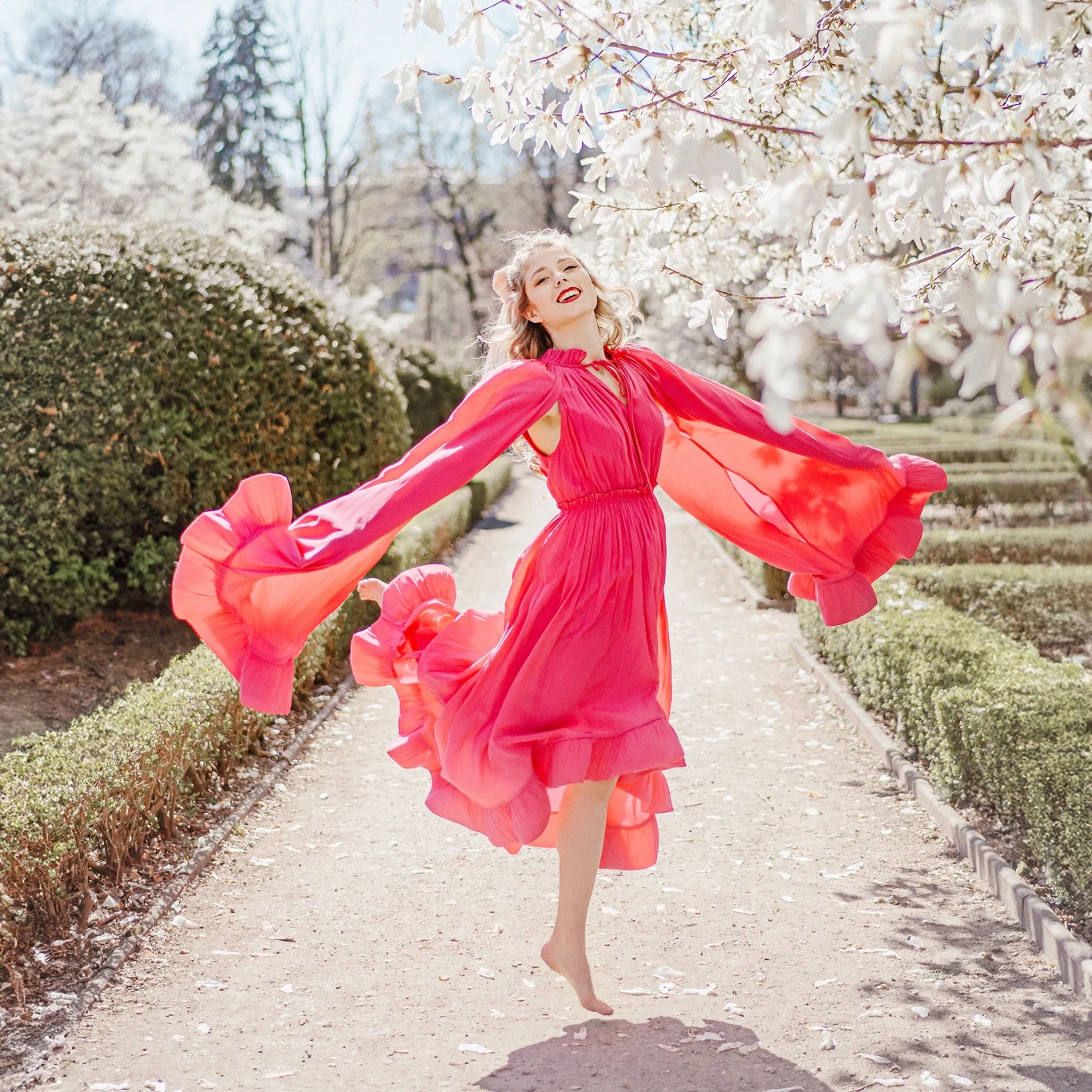 Karolina Lizer prezentuje piosenkę "Całuj mnie w nos" Kobieta w różowej, zwiewnej sukni skacze z radością wśród kwitnących białymi kwiatami drzew w parku, wokół alejki widać zielone żywopłoty i słoneczną pogodę.