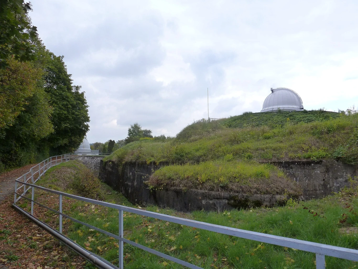 Dzieje fortu i sekrety jego budowy będzie można poznać podczas spaceru z przewodnikiem w czerwcu 2025 r. Obserwatorium astronomiczne z charakterystyczną kopułą na zielonym wzgórzu, otoczone bujną roślinnością oraz metalowym ogrodzeniem; ścieżka prowadzi wzdłuż rowu i lasu po lewej stronie.
