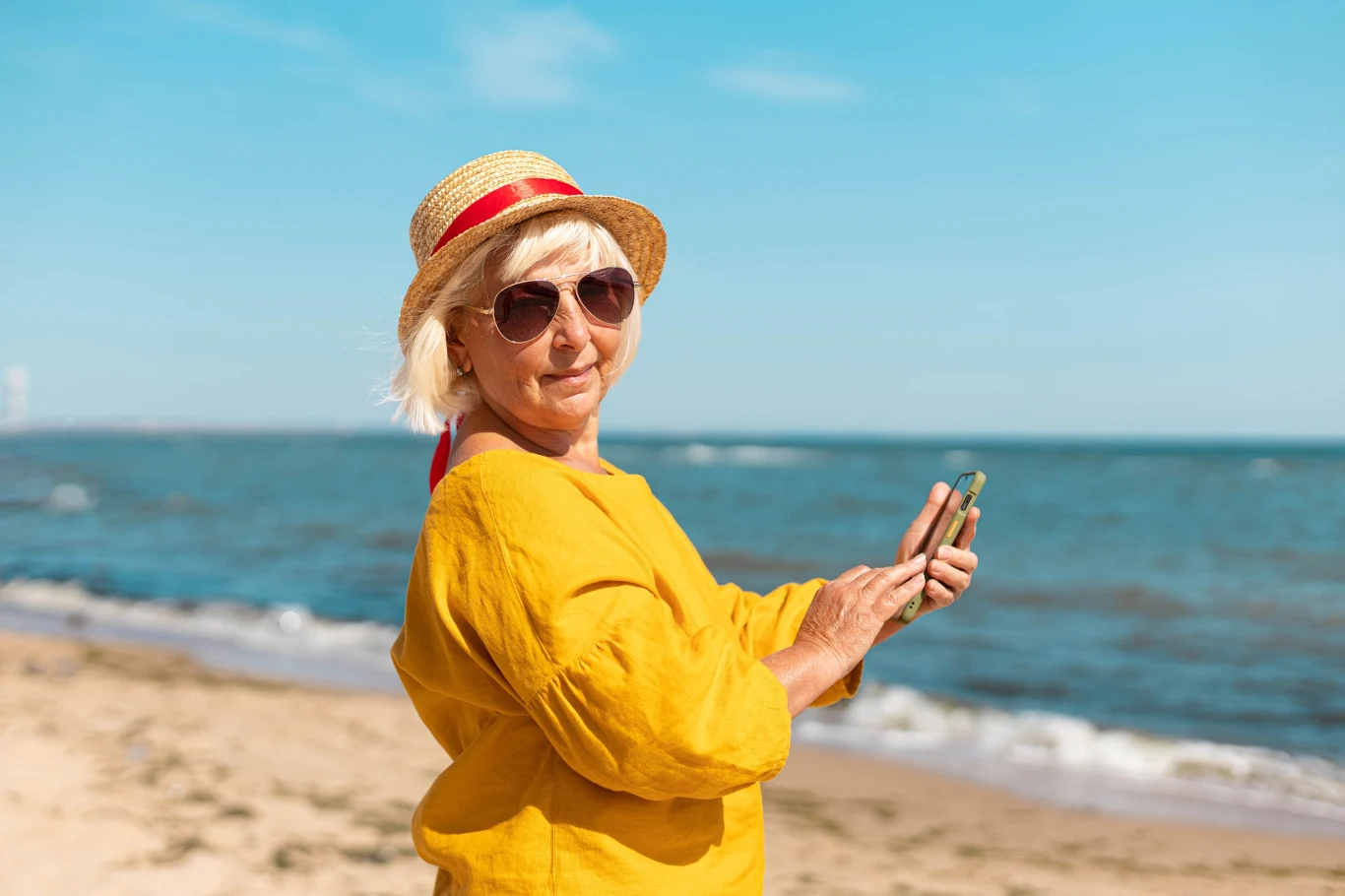 Starsza kobieta w okularach przeciwsłonecznych i słomkowym kapeluszu, ubrana w żółtą bluzkę, stoi na plaży, trzymając telefon komórkowy, w tle widać morze i błękitne niebo.