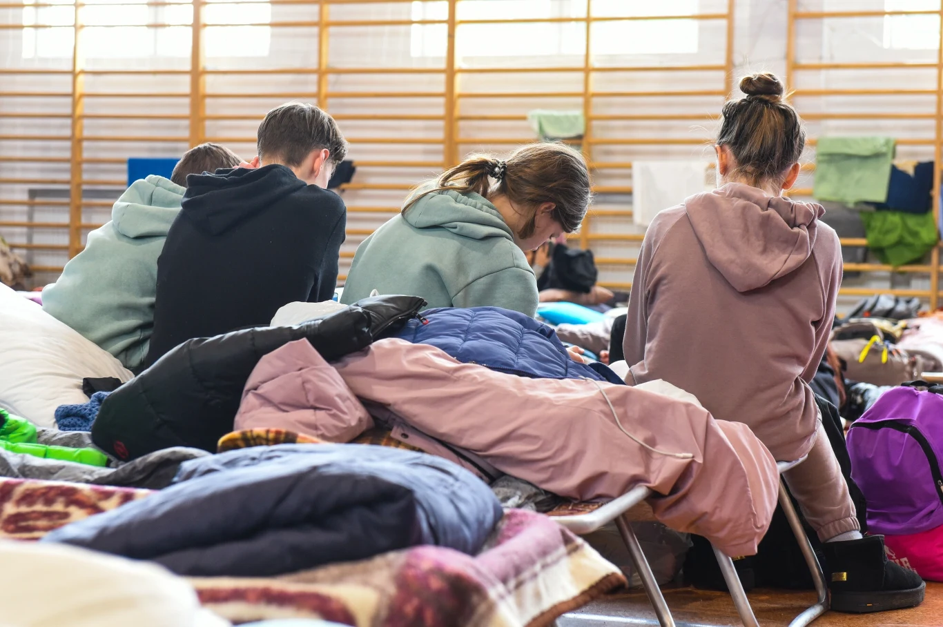 Czworo ludzi siedzących na łóżkach polowych, ubranych w codzienne ubrania, pochylonych do przodu, otoczonych walizkami, plecakami i rzeczami osobistymi, wnętrze przypomina salę sportową lub tymczasowe schronienie.