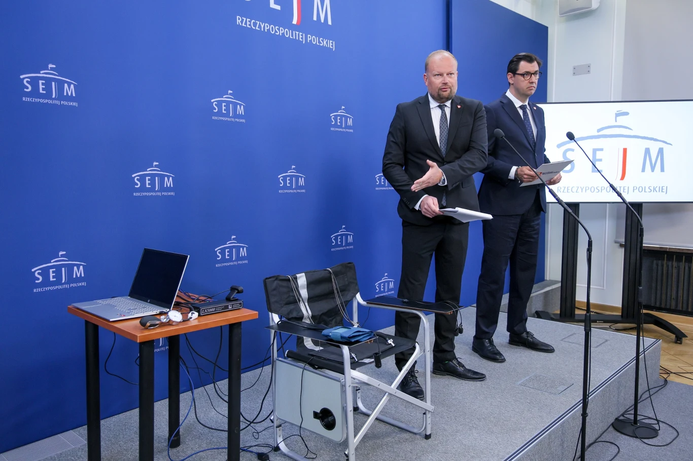 Politycy KO chcą przeprowadzić badanie Karola Nawrockiego wariografem Dwóch mężczyzn w garniturach stoi na tle niebieskiej ścianki z logotypami Sejmu RP, po prawej mikrofon i pulpit, po lewej krzesło, laptop i sprzęt elektroniczny na niewielkim stole.