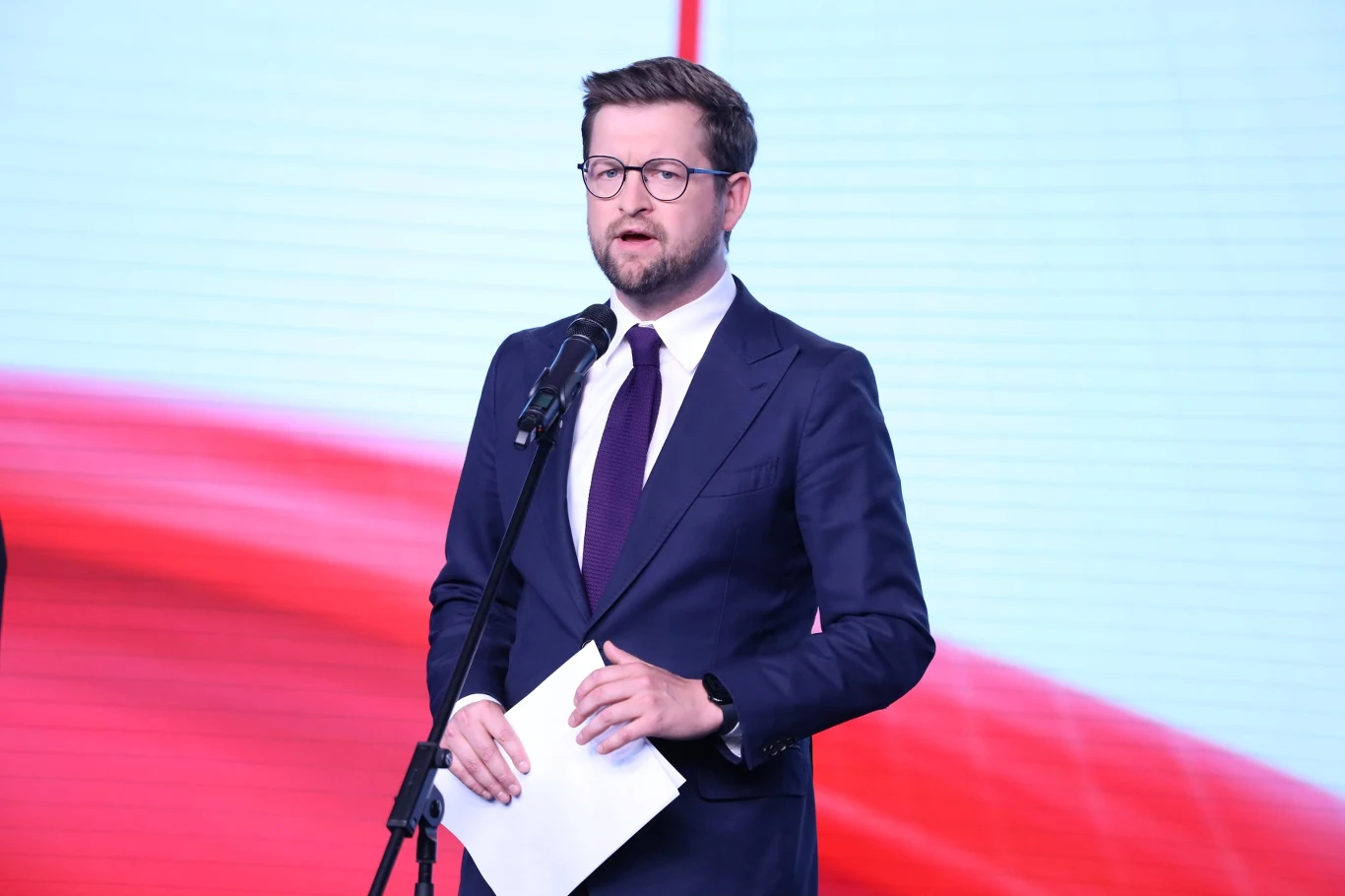 Poseł PiS Andrzej Śliwka na konferencji prasowej. Partia składa zawiadomienie ws. premiera i rzecznika MSWiA Mężczyzna w garniturze i okularach przemawia do mikrofonu, trzymając w ręku kartkę, na tle białoczerwonym z rozmytym motywem.