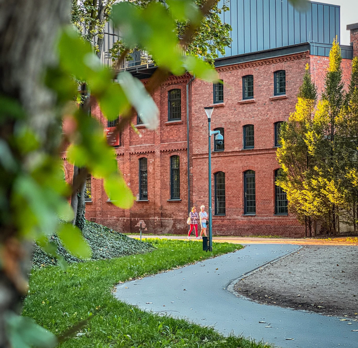 Ceglana, industrialna kamienica z dużymi oknami na tle zielonego skweru, dwie osoby idące chodnikiem oraz drzewa w ramie kadru.