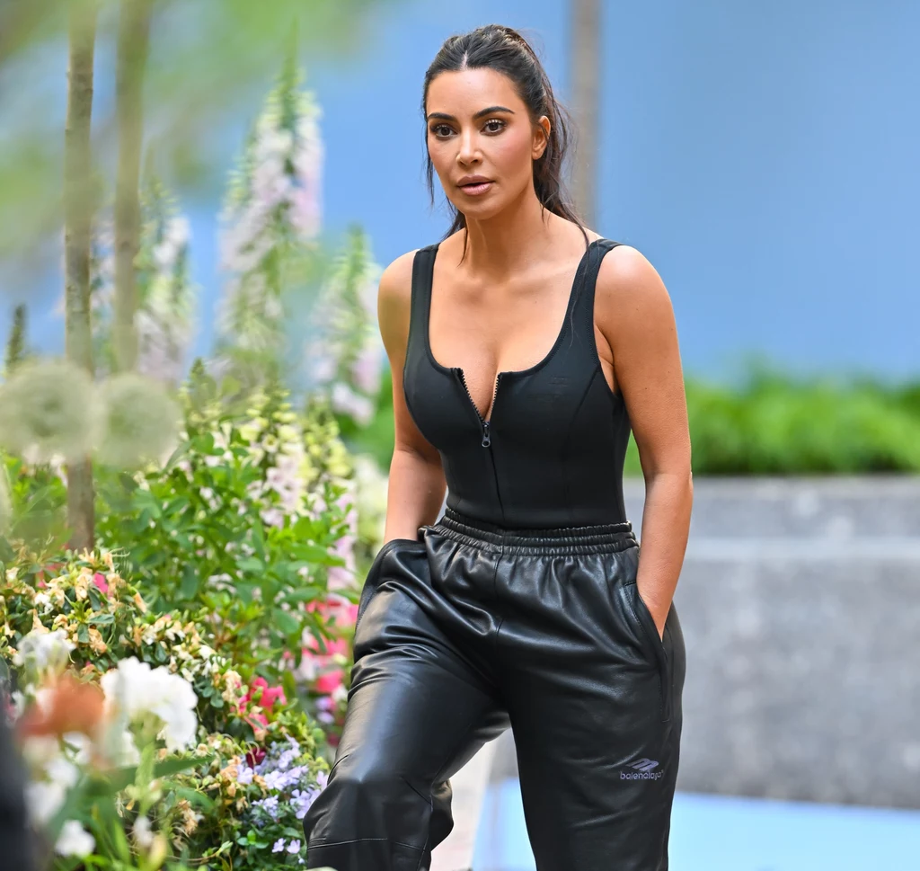 Czy Kim Kardashian porzuci życie gwiazdy, by stać się pełnoetatową prawniczką? Kobieta o długich ciemnych włosach ubrana w czarny top z zamkiem i skórzane spodnie stoi w ogrodzie pełnym kwiatów i zielonych roślin, w tle rozmyte pastelowe barwy oraz betonowy murek.