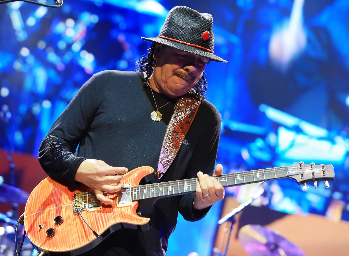 Carlos Santana zagra koncert w Polsce Mężczyzna w czarnym kapeluszu i czarnym ubraniu gra na gitarze elektrycznej na kolorowo oświetlonej scenie podczas koncertu, tło rozmyte z elementami scenografii i świateł.