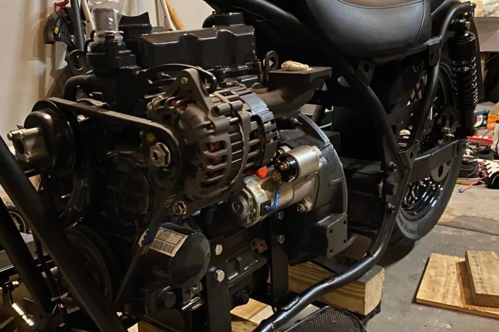 Silnik Diesla nie powala mocą, a dodatkowo jest cięższy i większy od jednostki benzynowej. Silnik zamontowany na ramie motocykla, widoczne szczegóły konstrukcyjne oraz elementy mechaniczne, całość ustawiona na drewnianych klockach na podłodze warsztatu.