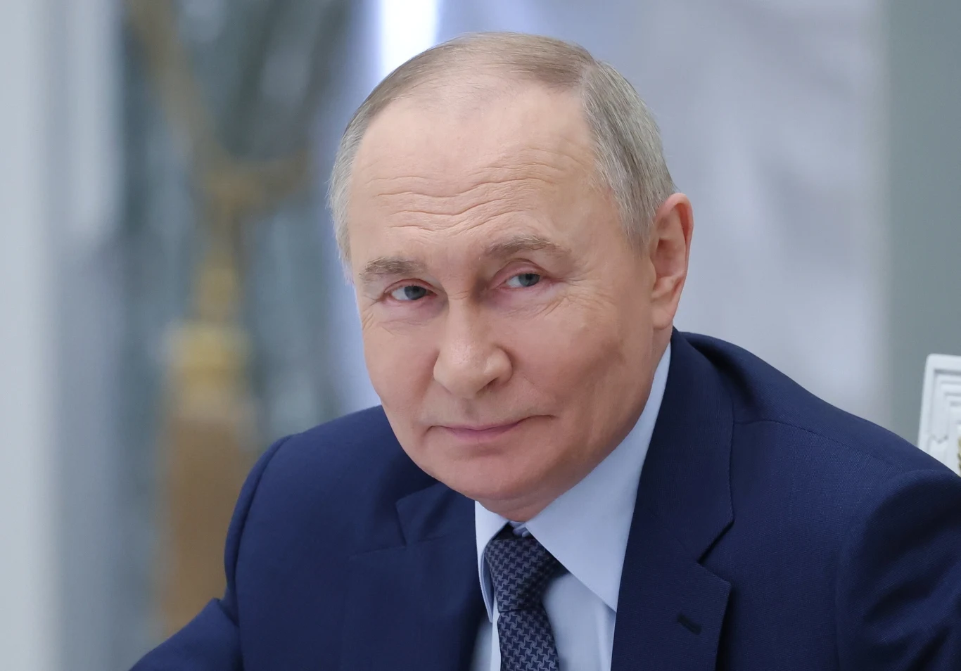 Władimir Putin Mężczyzna w średnim wieku w granatowym garniturze i jasnoniebieskiej koszuli siedzący przy stole, patrzący z lekkim uśmiechem w stronę aparatu, w tle zamazana jasna przestrzeń.