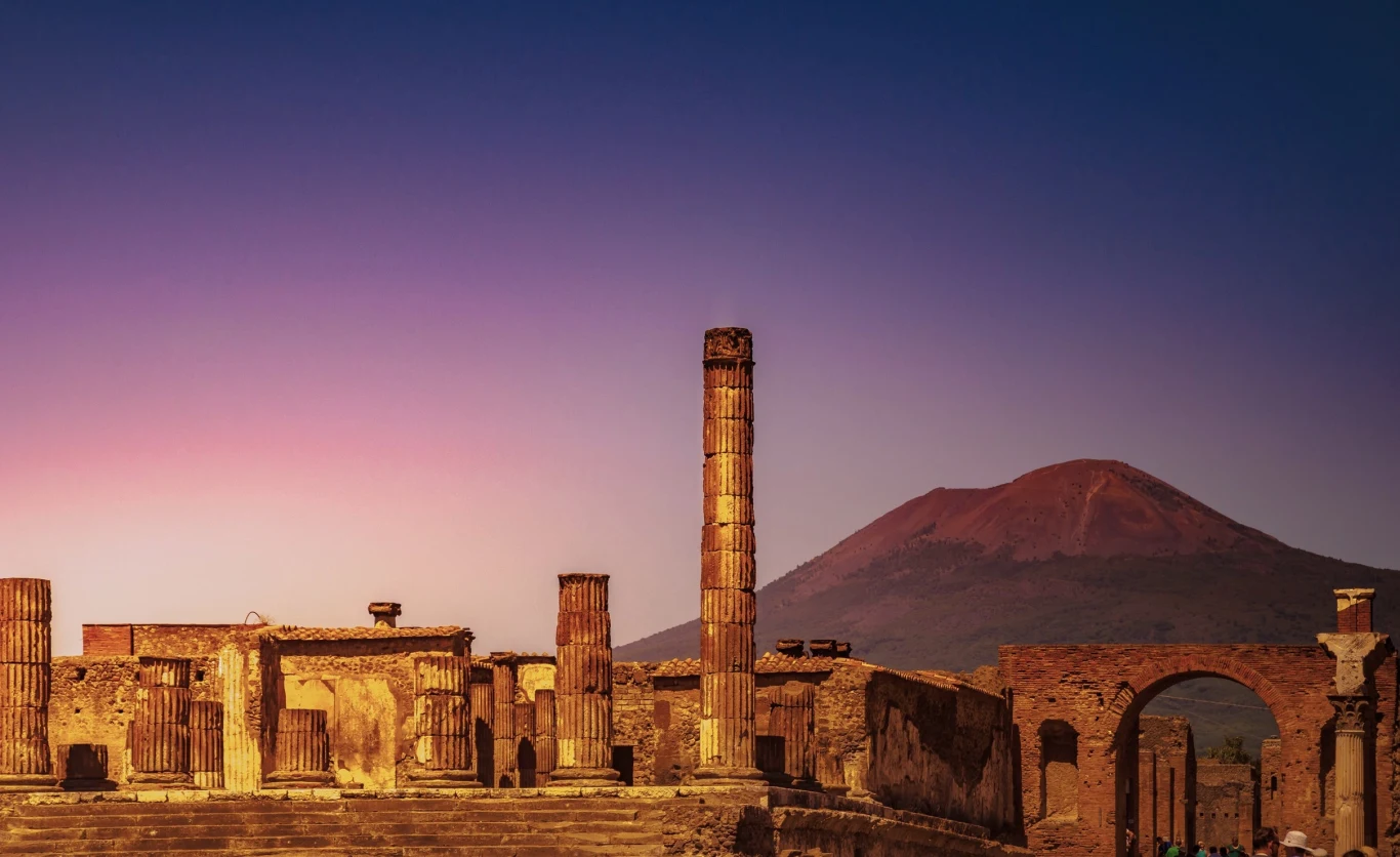 Znaleźli ślady życia w Pompejach po wybuchu Wezuwiusza Ruiny starożytnego miasta z dobrze zachowanymi kolumnami, ścianami i łukiem triumfalnym, w tle wulkan górujący nad krajobrazem na tle fioletowo-pomarańczowego nieba o zmierzchu.