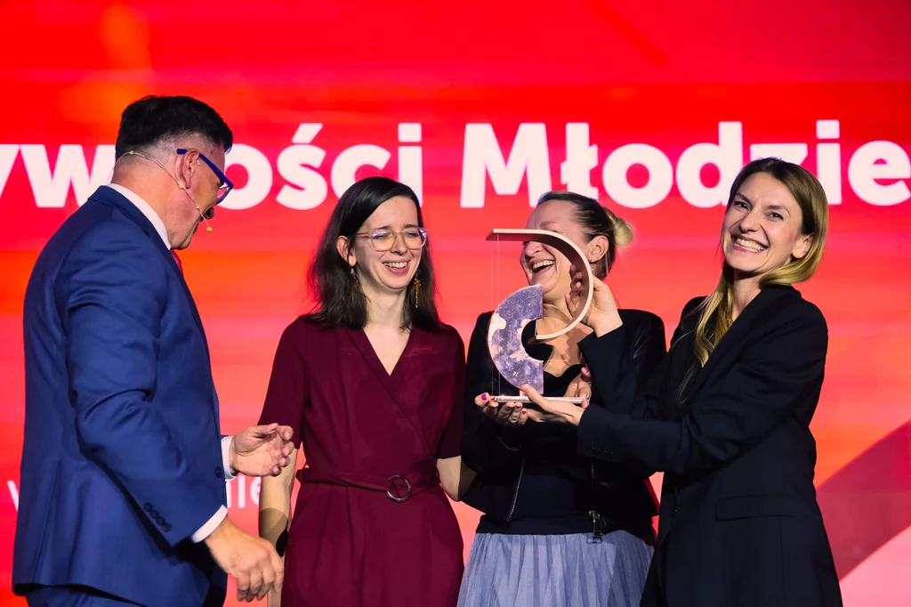 Nagroda Przewroty. Centrum Aktywności Młodzieży w Krośnie Czworo dorosłych osób w eleganckich ubraniach uśmiecha się i trzyma przezroczystą statuetkę, jedna z kobiet prezentuje ją dumnie, kolorowe, czerwone tło podkreśla uroczystą atmosferę wydarzenia.