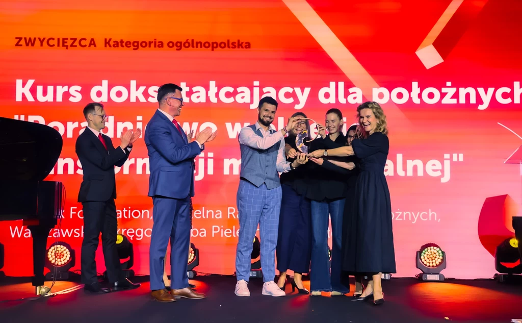 Kurs dokształcający dla położnych "Poradnictwo w okresie transformacji menopauzalnej" Grupa osób uśmiechniętych, ubranych elegancko, stojących na scenie podczas uroczystego wręczenia nagrody, za nimi jasny ekran z napisem wyróżniającym zwycięzców konkursu ogólnopolskiego dotyczącego kursu dla położnych.