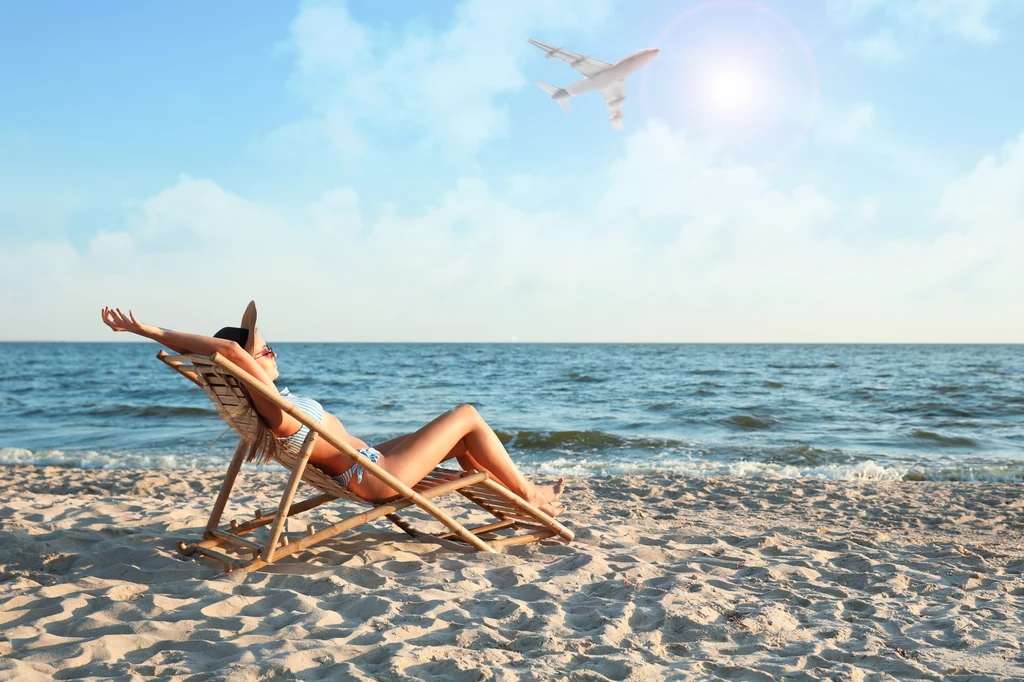 Sztuczki portali turystycznych Kobieta w bikini odpoczywa na leżaku na piaszczystej plaży, w tle spokojne morze, błękitne niebo oraz przelatujący samolot, słońce świeci na horyzoncie.