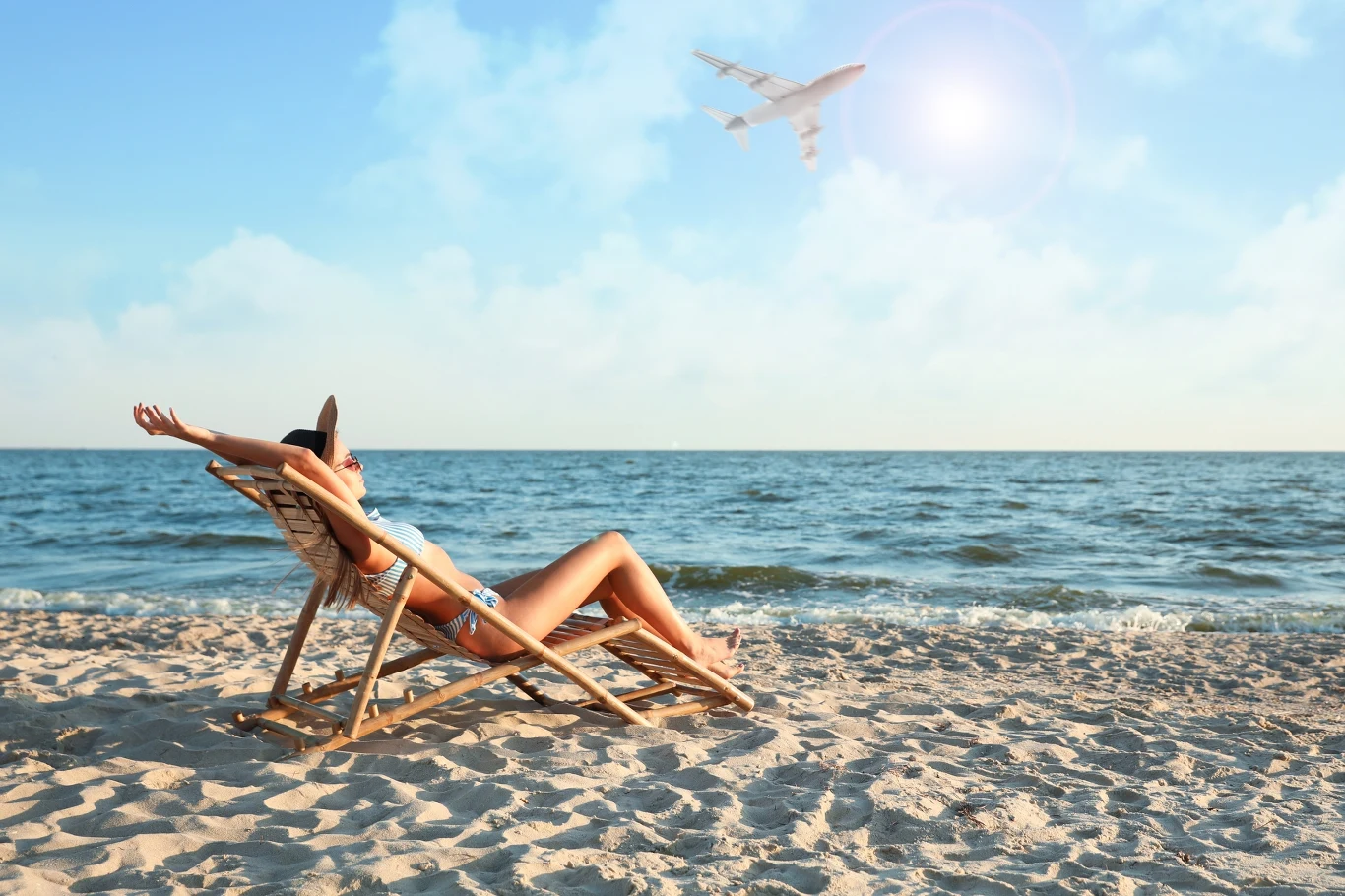 Kobieta w bikini odpoczywa na leżaku na piaszczystej plaży, w tle spokojne morze, błękitne niebo oraz przelatujący samolot, słońce świeci na horyzoncie.