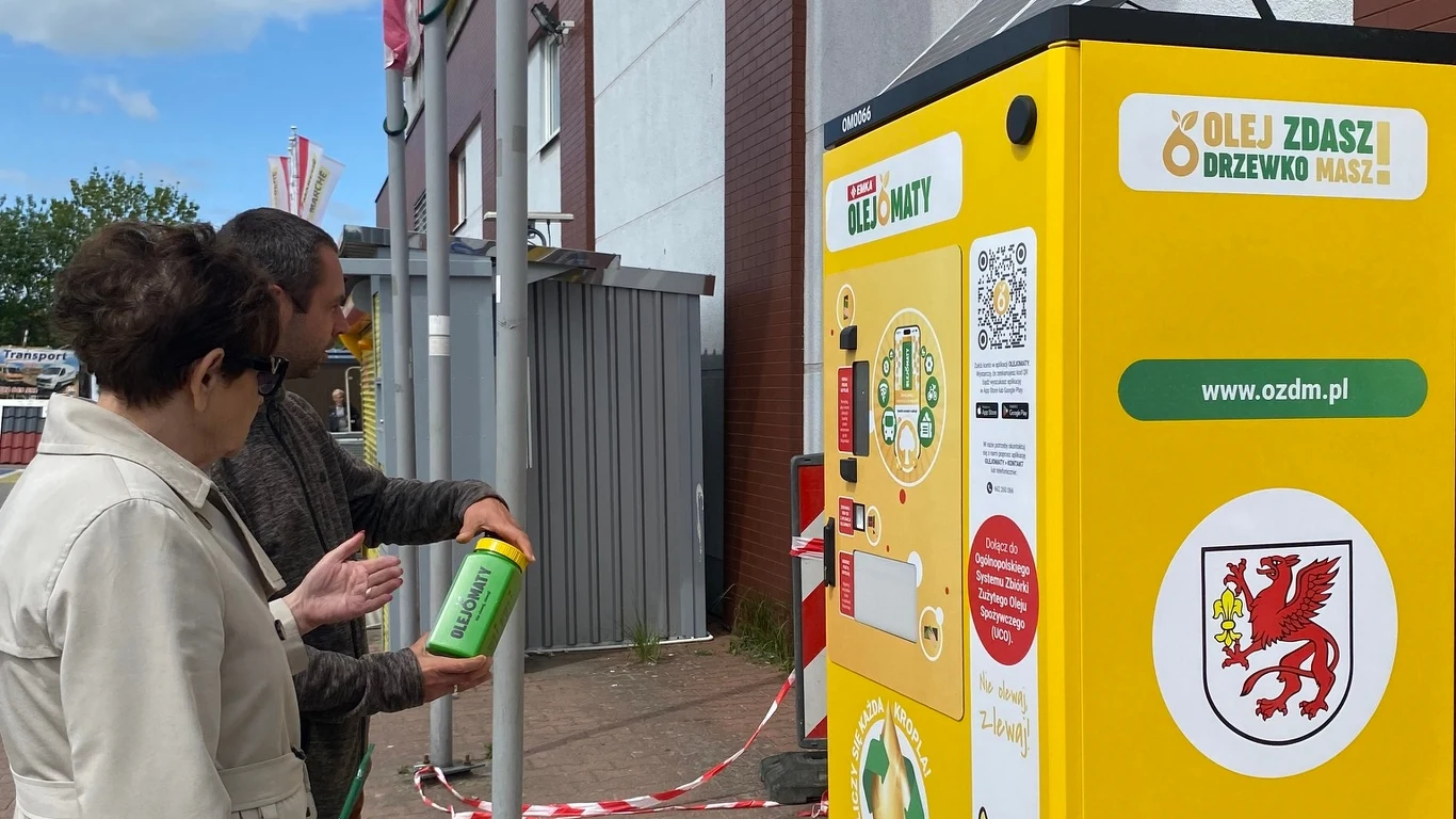 Olejomaty stanęły niedawno m.in. w Gryficach Para osób używa specjalnego automatu do zbierania zużytego oleju kuchennego, trzymając specjalny pojemnik przed kolorową maszyną z informacjami i logo.
