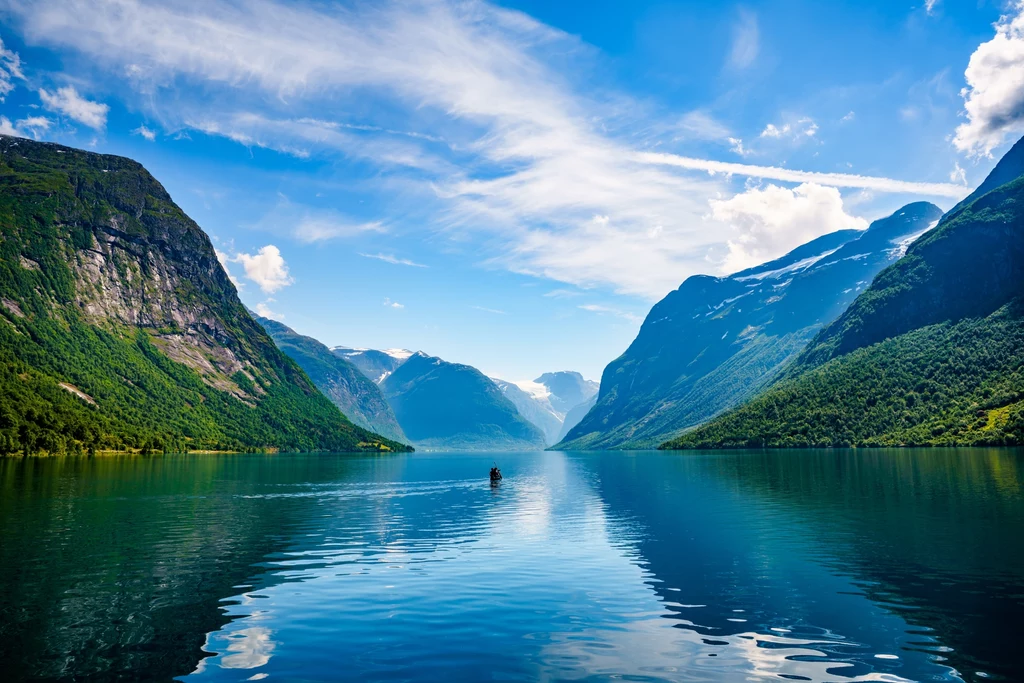 Norweskie fiordy to wspaniałe miejsce na wypoczynek w ciszy i spokoju Gładka tafla wody otoczona wysokimi, zielonymi wzgórzami i stromymi klifami, na środku niewielka łódka, nad całością rozpościera się błękitne niebo z delikatnymi chmurami.