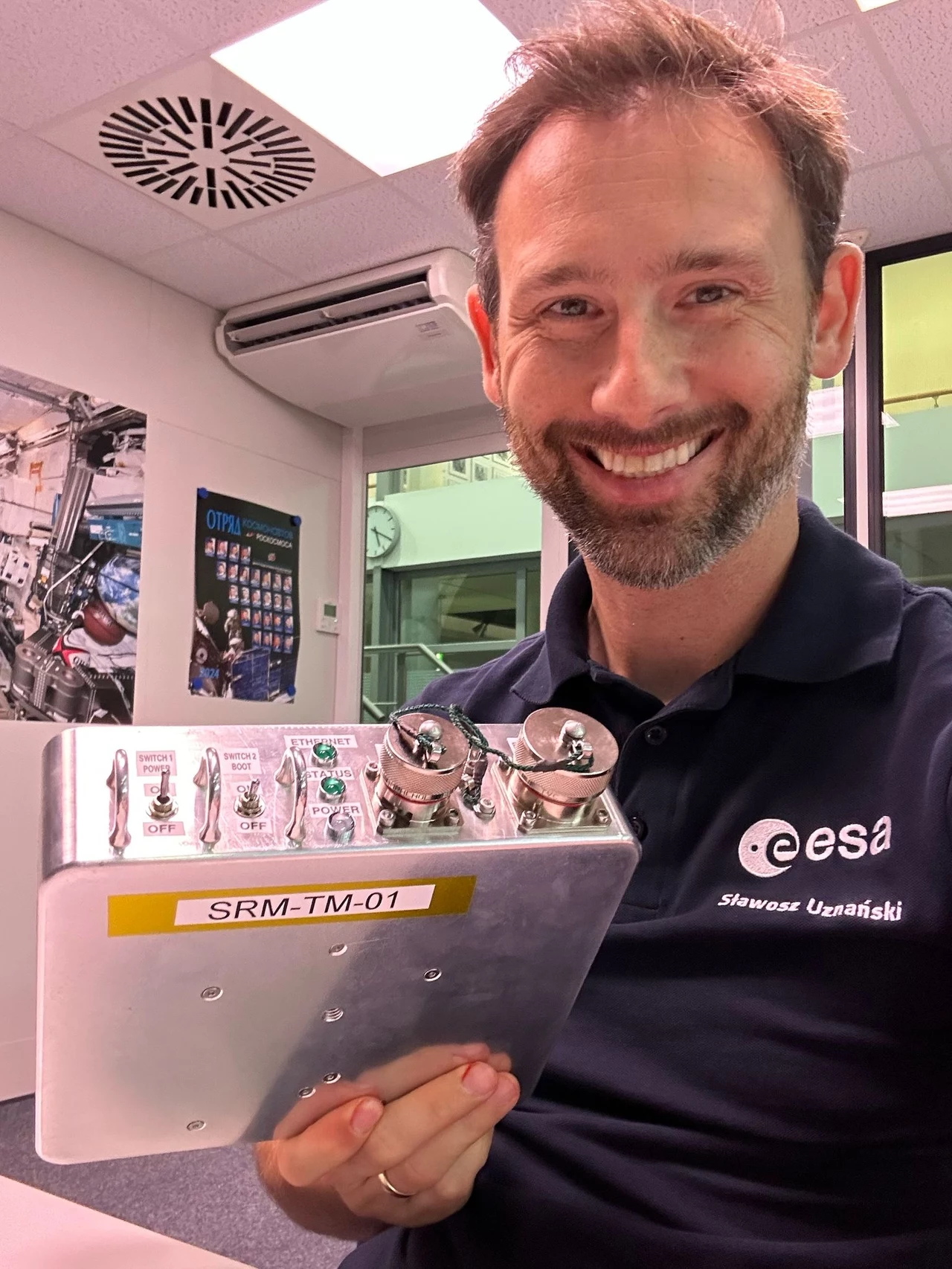 Dr Sławosz Uznański-Wiśniewki, z opracowanym w ramach eksperymentu RadMon-on-ISS instrumentem Scalable Radiation Monitor, podczas szkolenia na temat instalacji urządzenia w ESA European Astronaut Centre (EAC) w Kolonii w Niemczech. Mężczyzna w koszulce z logo ESA trzyma w rękach urządzenie oznaczone jako SRM-TM-01, uśmiecha się do aparatu, w tle widać nowoczesne biuro z plakatami o tematyce kosmicznej.
