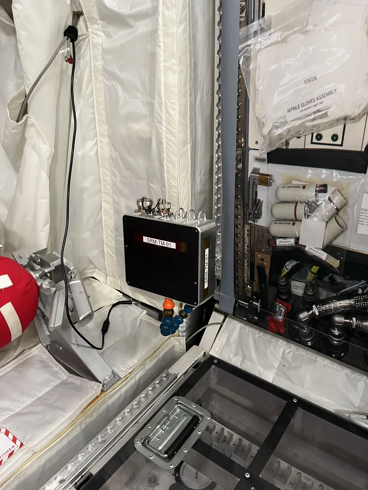Scalable Radiation Monitor (SRM), instrument opracowany w ramach eksperymentu RadMon-on-ISS, zainstalowany w module treningowym Columbus w ESA European Astronaut Centre (EAC) w Kolonii w Niemczech. Panel kontrolny umieszczony na ścianie, wokół niego specjalistyczny sprzęt techniczny, białe pokrowce ochronne, narzędzia i elementy wyposażenia wnętrza laboratoryjnego lub pojazdu kosmicznego.