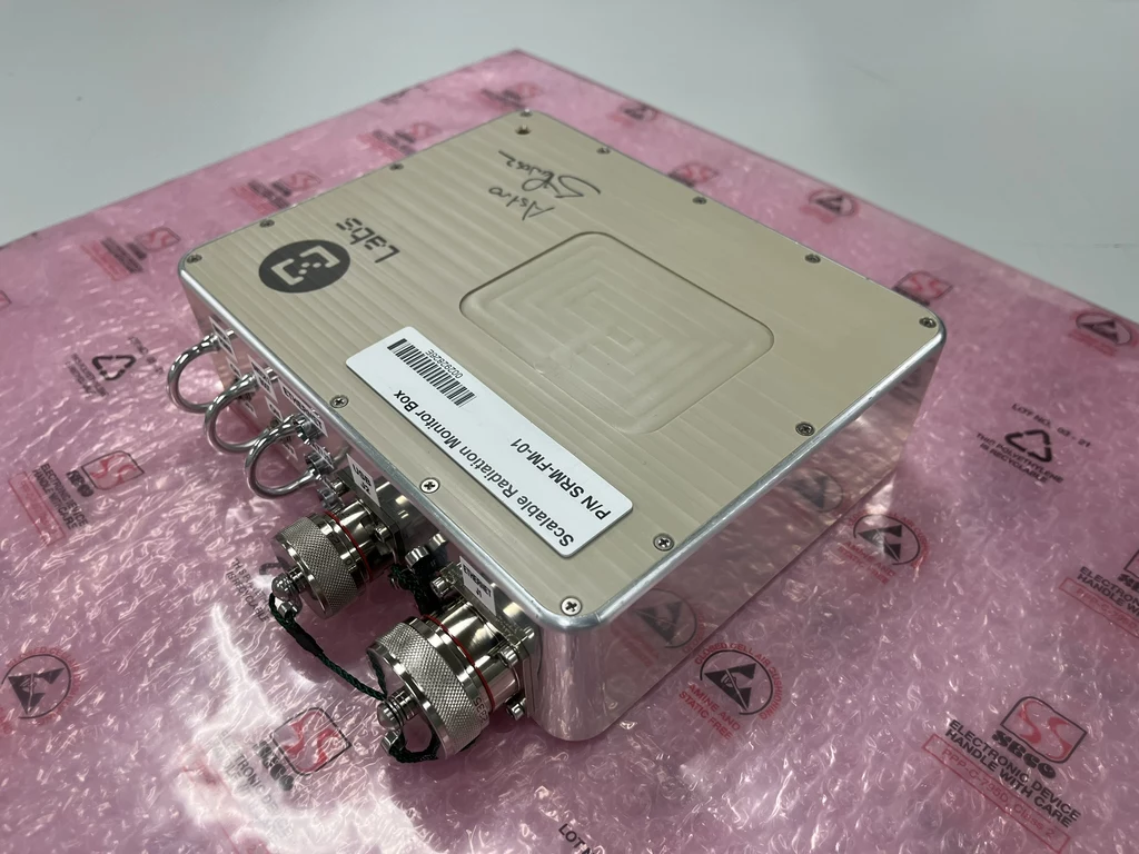 Opracowany przez zespół SigmaLabs, w ramach eksperymentu RadMon-on-ISS, instrument Scalable Radiation Monitor. Metalowa obudowa urządzenia elektronicznego wyposażona w złącza i naklejki identyfikacyjne, leżąca na różowej macie antystatycznej.