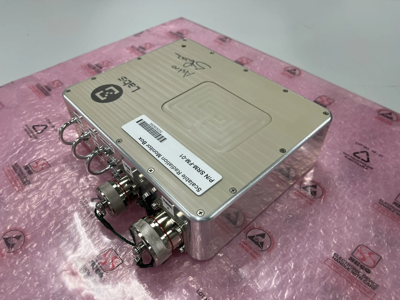 Metalowa obudowa urządzenia elektronicznego wyposażona w złącza i naklejki identyfikacyjne, leżąca na różowej macie antystatycznej.