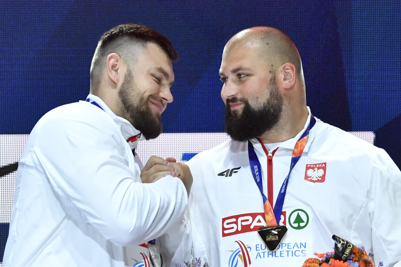 Konrad Bukowiecki (z lewej) i Michał Haratyk Dwóch sportowców w białych dresach z medalami na szyi i biało-czerwonymi emblematami, stojących obok siebie i uśmiechających się, jeden z nich trzyma medal, na szyjach widoczne niebieskie wstążki, w rękach bukiet kwiatów.