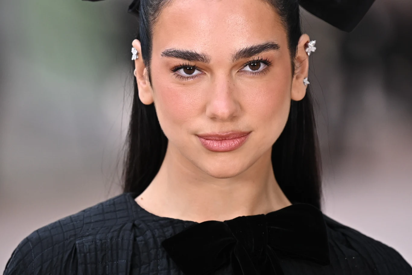 Dua Lipa Młoda kobieta z ciemnymi, gładko uczesanymi włosami spiętymi z tyłu, duże czarne kokardy po bokach głowy, delikatny makijaż, niewielkie kolczyki w uszach, ubrana w czarną bluzkę z dużą kokardą pod szyją, patrzy prosto w obiektyw z lekkim uśmiechem.