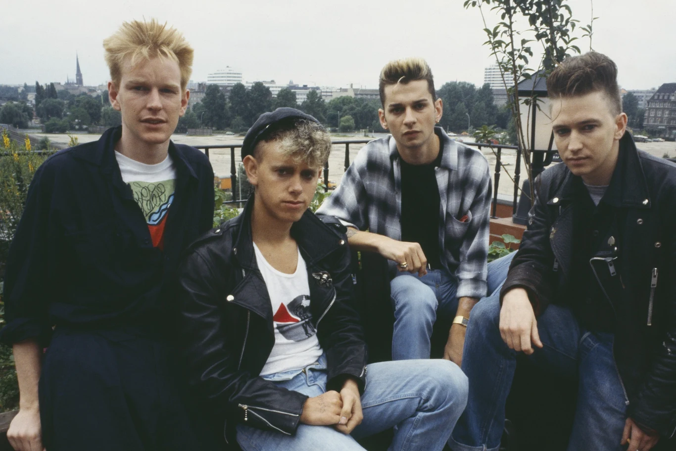 Depeche Mode w latach 80. Czterech młodych mężczyzn w stylizacjach charakterystycznych dla lat 80., pozujących na tle miejskiego krajobrazu z widoczną zielenią i budynkami w oddali.