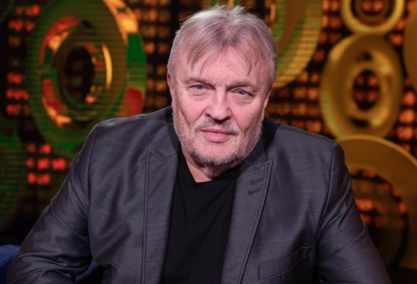 Krzysztof Cugowski skończył 75 lat Mężczyzna w średnim wieku w szarej marynarce i czarnej koszulce siedzi na tle abstrakcyjnych, złotych okręgów na ciemnym tle.