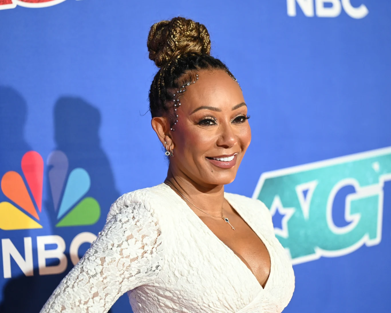 Mel B obchodzi swoje 50. urodziny Uśmiechnięta kobieta w białej, koronkowej sukience z upiętymi włosami w artystyczny kok stoi na tle niebieskiej ścianki z logotypami NBC oraz programu telewizyjnego.