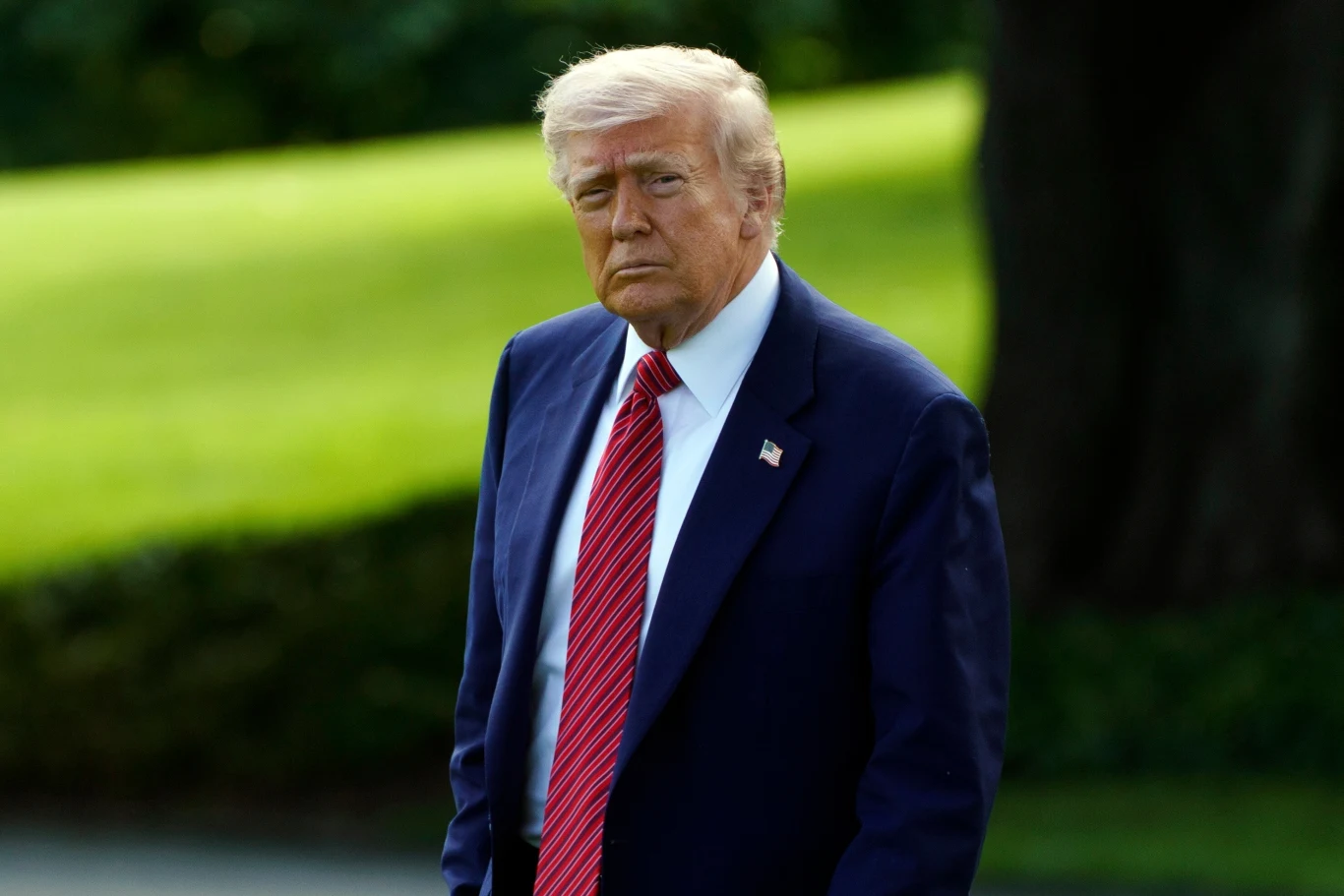 Donald Trump. Mężczyzna w garniturze i czerwonym krawacie spaceruje na świeżym powietrzu, w tle widoczne zielone drzewa i trawnik.
