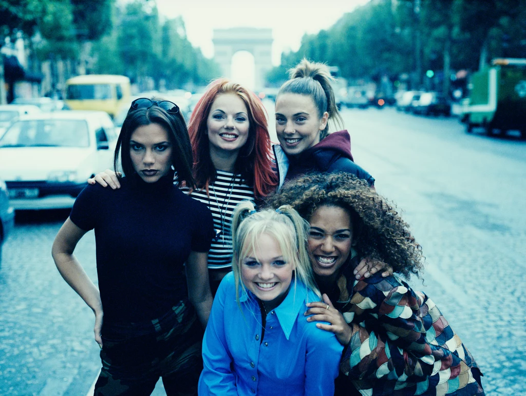 Spice Girls w 1996 r. Pięć młodych kobiet pozujących razem na szerokiej ulicy, w tle widoczny Łuk Triumfalny i ruch uliczny; dziewczyny są uśmiechnięte, kolorowo ubrane, stanowią główny punkt kadru.