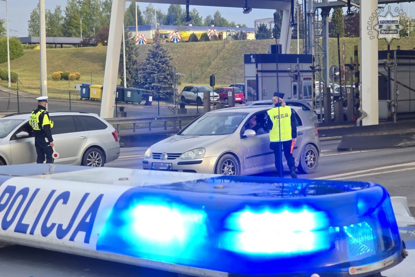 Stróże prawa zatrzymali 4 osoby w stanie nietrzeźwości i 3 w stanie po użyciu alkoholu. Policjant kieruje ruchem obok zatrzymanego srebrnego samochodu osobowego przy punkcie kontrolnym, na pierwszym planie widoczne niebieskie światła radiowozu policyjnego, w tle stacja tankowania i zieleń miejska.