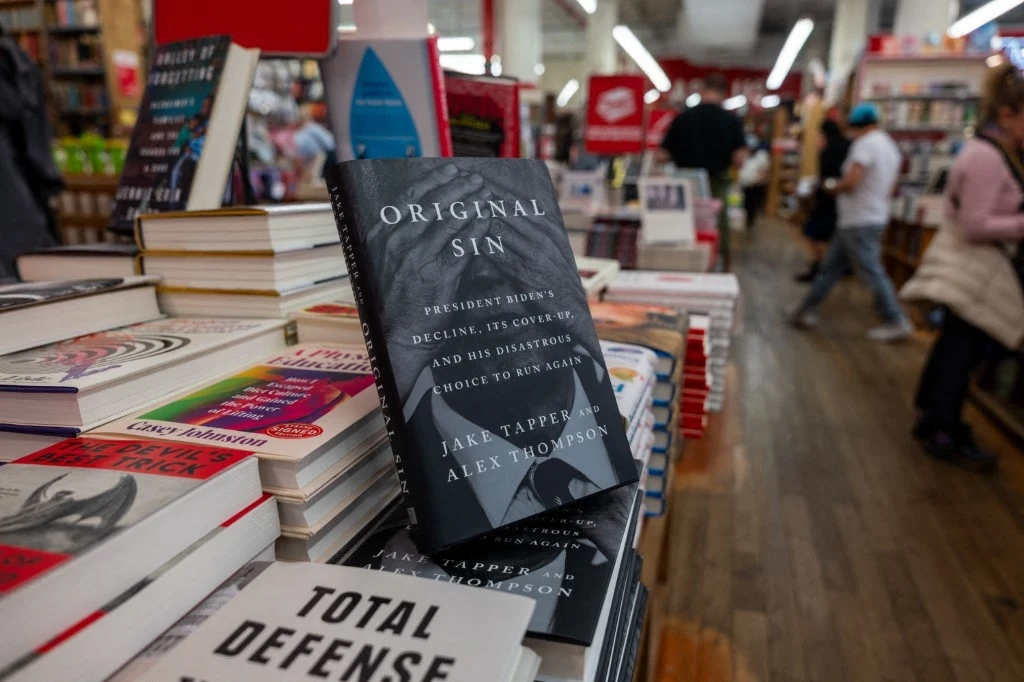 Stosy książek wystawione na półkach księgarni, na pierwszym planie widoczna książka o ciemnej okładce pt. 'Original Sin', obok inne tytuły o tematyce politycznej, w tle ludzie przeglądający książki.