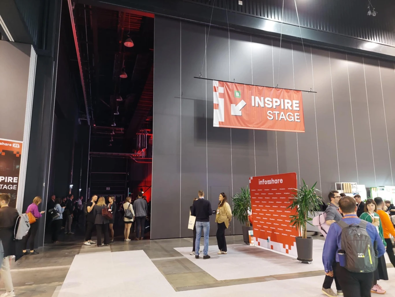 Uczestnicy konferencji gromadzą się przy wejściu na Inspire Stage, w tle znajduje się duży baner z nazwą sceny oraz ścianka do zdjęć z logotypem wydarzenia, po bokach widoczne rośliny i część przestrzeni hali.