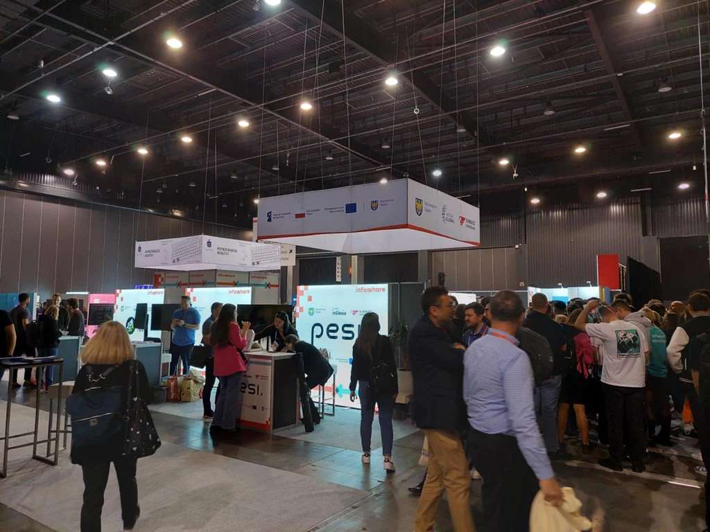 Centralną część hali Amber Expo zajmowały dziesiątki stoisk firm technologicznych Tłum ludzi zwiedzających stoiska wystawiennicze podczas wydarzenia branżowego w dużej, oświetlonej hali z licznymi banerami promocyjnymi i stanowiskami firm.
