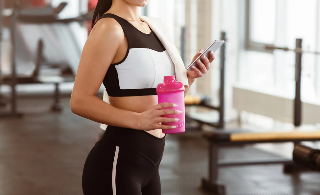 Coraz więcej osób szuka wsparcia regularnych treningów Kobieta w stroju sportowym trzymająca różowy shaker oraz telefon w jasnej, nowoczesnej siłowni, z ręcznikiem zarzuconym na ramię.