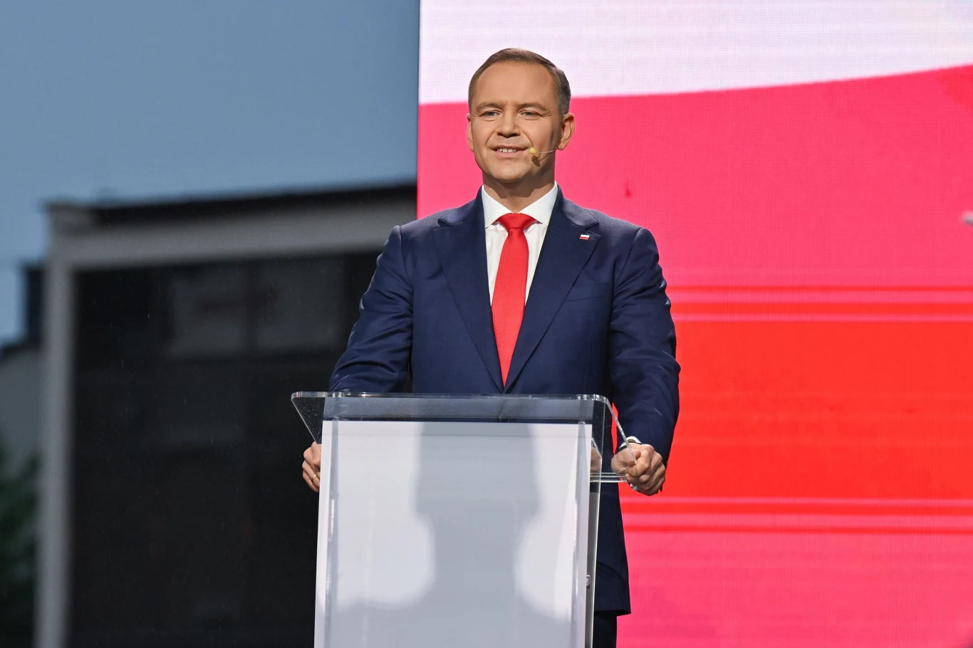 Karol Nawrocki na scenie w Końskich Mężczyzna w granatowym garniturze z czerwoną krawatą stoi za przezroczystą mównicą na tle biało-czerwonej planszy, sprawiając wrażenie, że przemawia publicznie podczas oficjalnego wydarzenia.