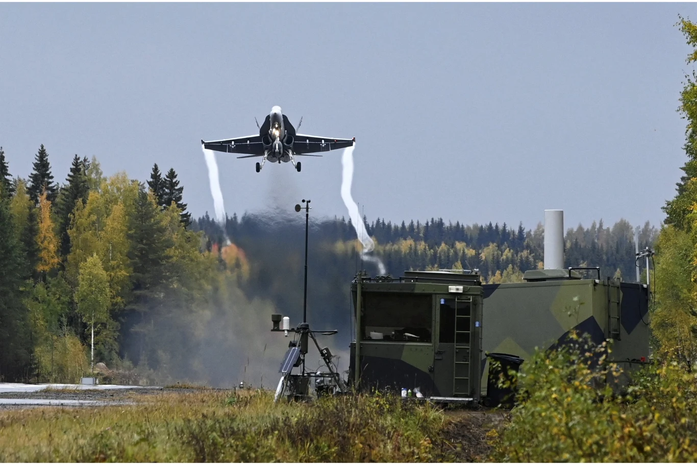 Myśliwiec wojskowy wznosi się pionowo do góry, za nim widoczne są smugi spalin, w tle gęsty las iglasty, a na pierwszym planie wojskowy kontener i sprzęt elektroniczny camuflowany wśród roślinności.