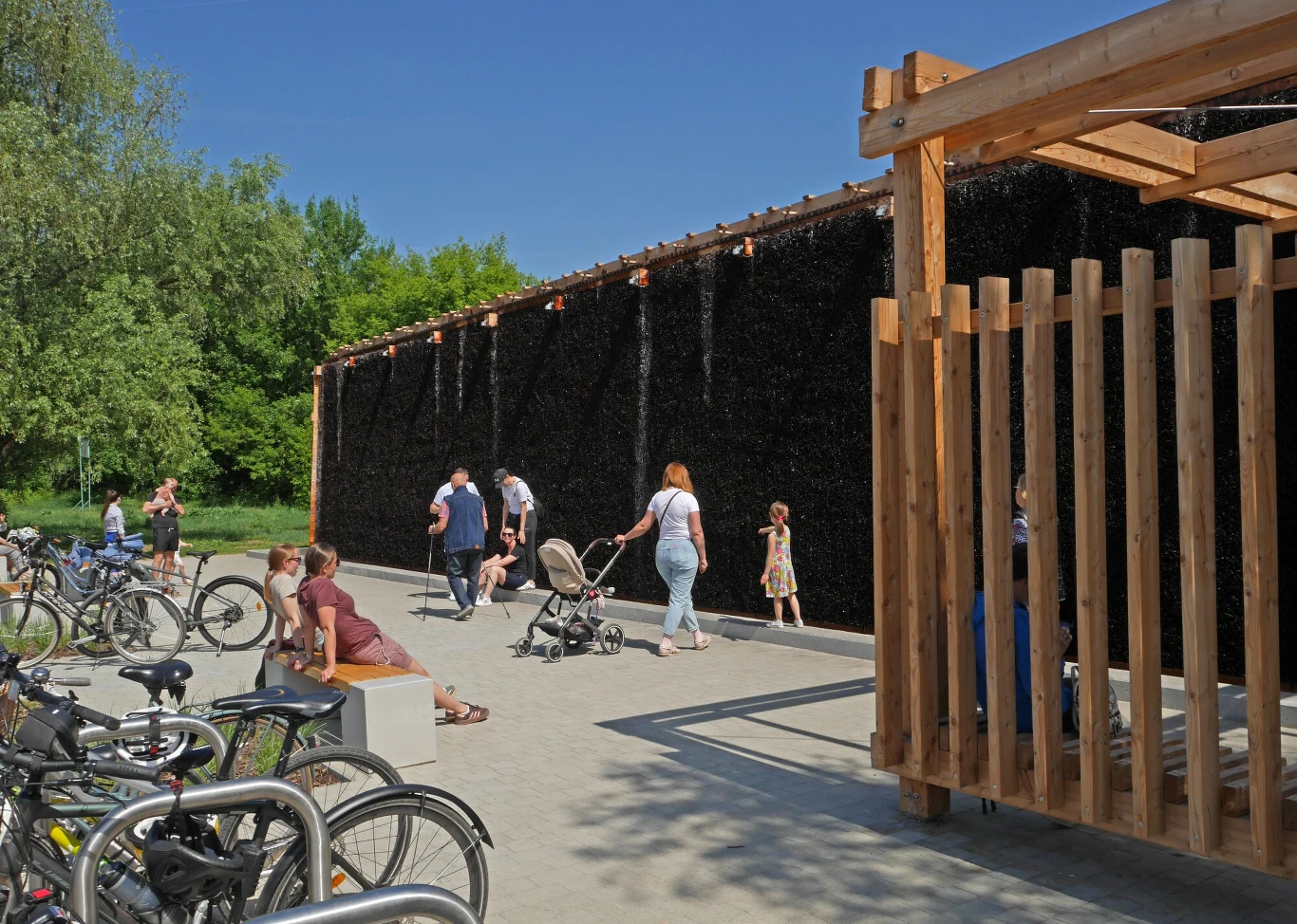 Naukowcy odkryli, że tężnie mogą być siedliskiem groźnych patogenów Uzdrowiskowa tężnia solankowa otoczona cieniem, przy której odpoczywają i przechodzą osoby dorosłe oraz dzieci, w otoczeniu rowerów i zielonych drzew, z drewnianą konstrukcją wejściową po prawej stronie.